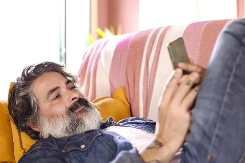 Happy smiling man using a smartphone device while sitting on a sofa at home.  Stock Photos