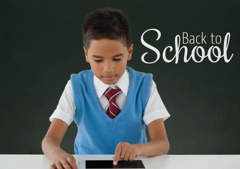 Happy student boy at table using a tablet against green blackboard with back to Foto stock