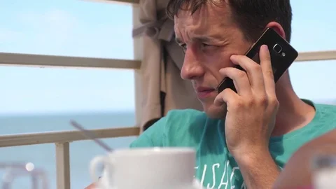Happy successful man drinking coffee in beach cafe and talking on cellphone by 스톡 동영상 83553264