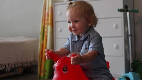 A happy toddler playing on a red rubber bouncy horse toy in a bright and cozy Stock Footage 345611526