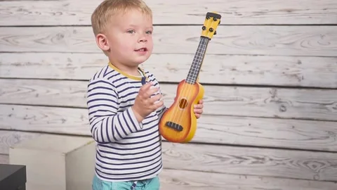 Happy two year old boy plays his guitar or ukulele and sings songs, sitting in Video stock 143095942