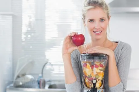 Happy woman leaning on her juicer full of fruit and holding red apple Stock Photos