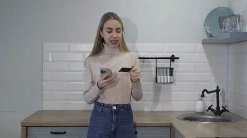 Happy woman makes a purchase using a credit card and a smartphone in the kitchen Stock Footage 260463569