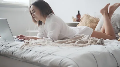 Happy woman is using laptop while lying in bed and smiling. girl in a man's Stock Footage 125978393