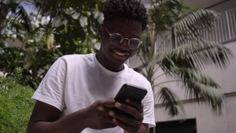 Happy young African using cell phone sitting with palm trees in background Stock Footage 245453284