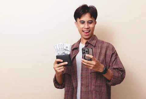 Happy young Asian man using smartphone while holding wallet full of money w.. Stock Photos