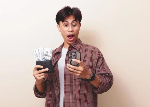 Happy young Asian man using smartphone and holding wallet full of money wit.. Stock Photos