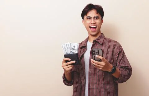 Happy young Asian man using smartphone while holding wallet full of money w.. Stock Photos