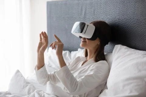 Happy young brunette woman resting in bed, wearing VR googles. Stock Photos