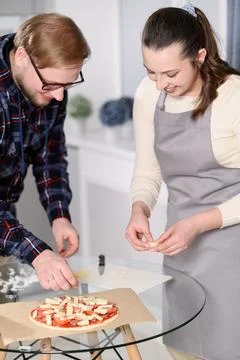 Happy young couple smiling, adding cheese toppings to homemade pizza in modern Stockfoto's