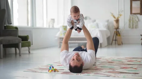 Happy young dad lying on floor carpet, lifting in air adorable little baby son Stock Footage 153414077