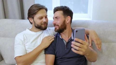 Happy young gay couple using mobile phone for video call while sitting on sof Stock Footage 169730484