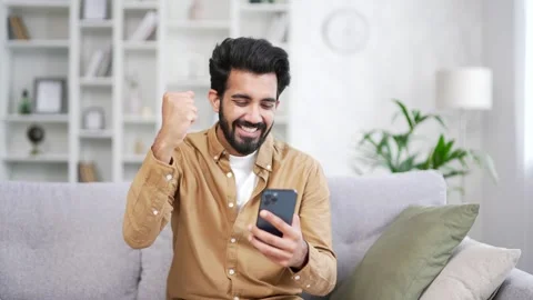 Happy young handsome man received great news on smartphone while sitting on sofa Stock Footage 252201652