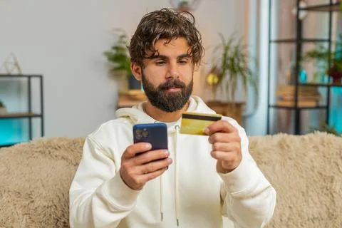 Happy young Hispanic man guy using credit bank card and cellphone while shopping Stock Photos