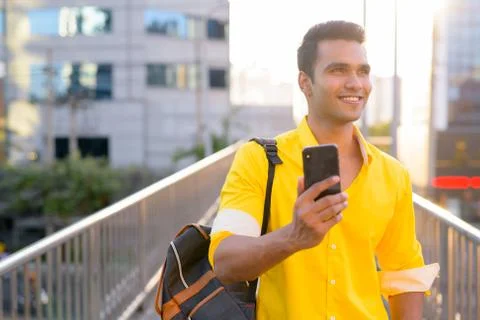Happy young Indian man with backpack thinking while using phone on the Foto stock