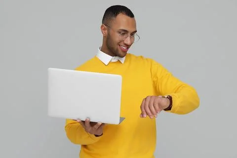 Happy young intern with laptop checking time on light grey background Stock Photos