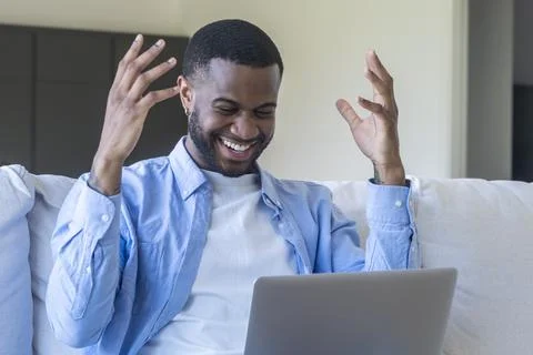 Happy young man celebrating while using laptop on couch at home, smiling an.. Foto stock