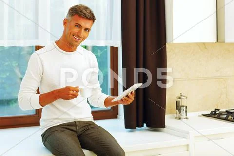 Happy young man at home drinking coffee and reading news on his electronic ta Stock Photos