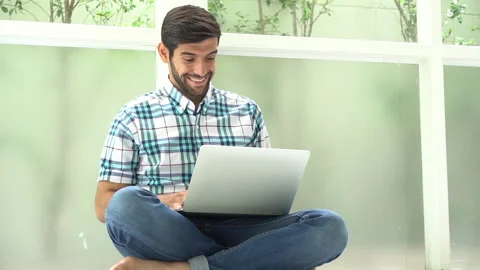Happy young man sitting on floor working with laptop computer on window at ho Stock Footage 134398947