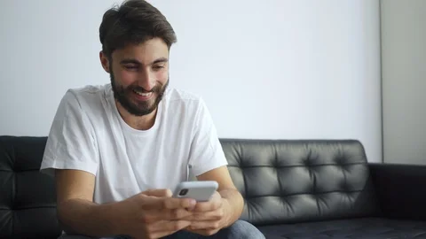 Happy young man sitting on sofa typing message on phone at home Video stock 119309076