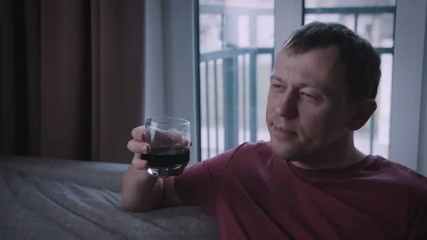 A happy young man is sitting on a sofa, smiling and drinking a beverage from a Stock Footage 316956948