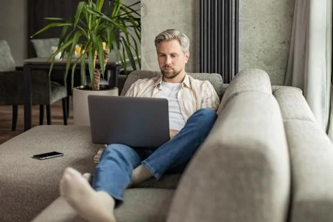 Happy young man sitting on sofa and working on laptop from home. Stock Photos