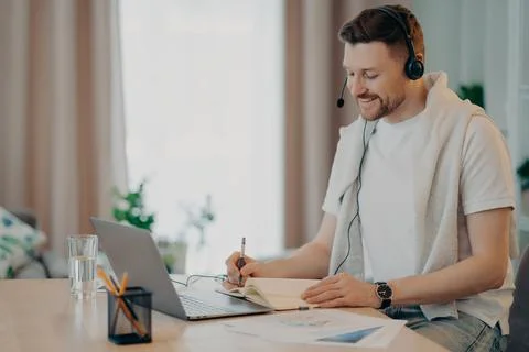 Happy young man sitting at the table at home and making notes in note book Stock Photos