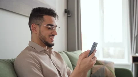 Happy young man smiling looking at cell phone sitting in living room at home Stock Footage 170679351