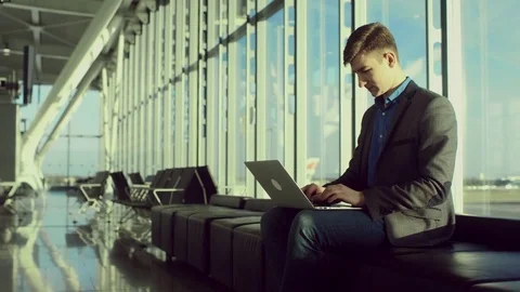 Happy young man is typing something on his laptop while sitting in airport Stock Footage 77602630