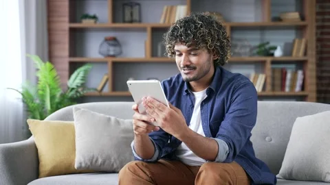 Happy young man is using a digital tablet while sitting on sofa in living room  库存影片 297871898