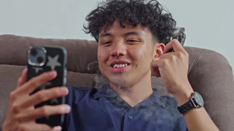 Happy young man using smartphone while vaping on sofa Stock Footage 332485733