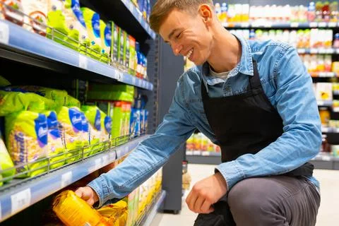 Happy young manager in apron man doing stocktaking of cereals on shelves sitting Stock Photos