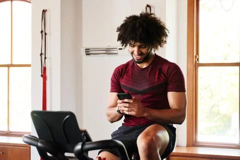Happy young middle eastern man using mobile phone on exercise bike at the gym Stock Photos