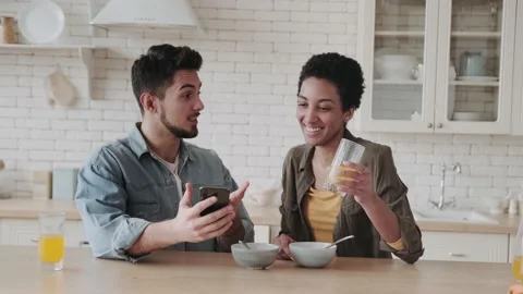 Happy young multiethnic couple looking at smartphone and smiling at breakfast in Video stock 236486592