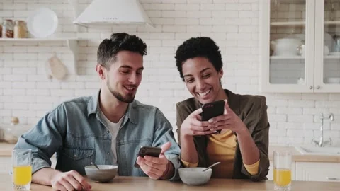 Happy young multiethnic couple looking at smartphone and smiling at breakfast in Stock Footage 236486708