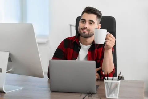 Happy young programmer drinking tea while working with laptop in office Stock Photos