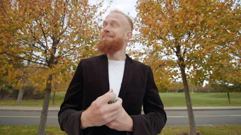 Happy young red-haired man counting cash standing alone on alley in autumn park Stock Footage 256045677