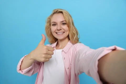 Happy young woman showing thumb up gesture while taking selfie on light blu.. Stock Photos