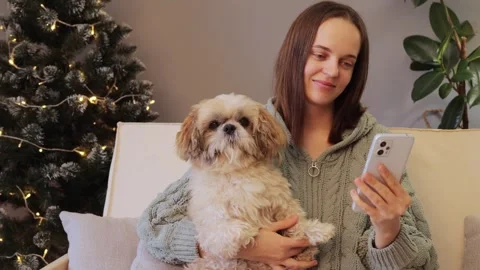 Happy young woman using smart phone in front of Christmas tree at home while Stock Footage 255110133