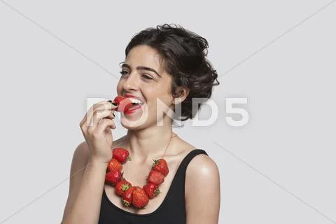 Happy young woman wearing strawberry necklace as she eats one piece over gray Stock Photos