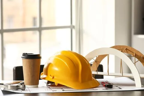 Hardhat with coffee cup, bridge model and house plan on desk in office Stock Photos