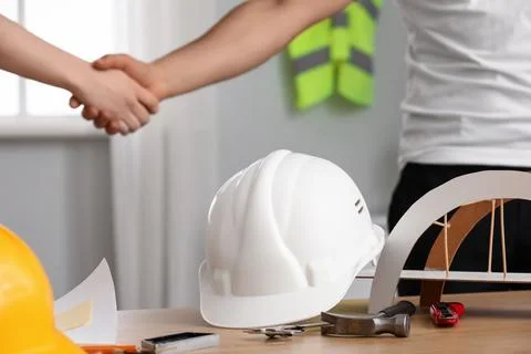 Hardhat with tools on desk against construction workers shaking hands in offi Stock Photos