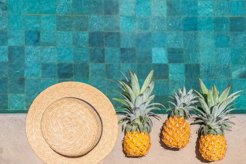 A hat and three pineapples are in a row by the water. Top view, copy space. Stock Photos