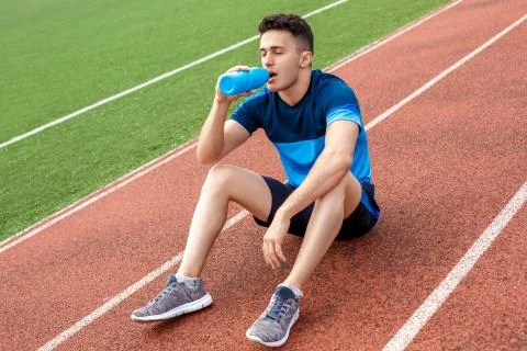 Having break. Young man on stadium outside sitting drinking water thirsty closed Stock Photos