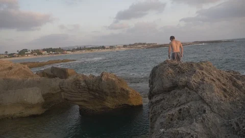 HD slow motion young man jumping from high rocky ledge Into the ocean. Stock Footage 116060159