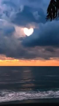 Heart shape appears in clouds over ocean during sunset near shore with wave.. Stock Photos