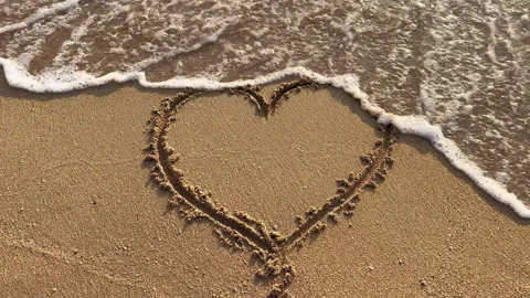 Heart shape drawn in sand on the beach with ocean waves washing over Stock Footage 282103457