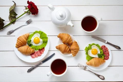 Heart shaped fried eggs, salad, croissants, salami sausage, rose flower Stock Photos