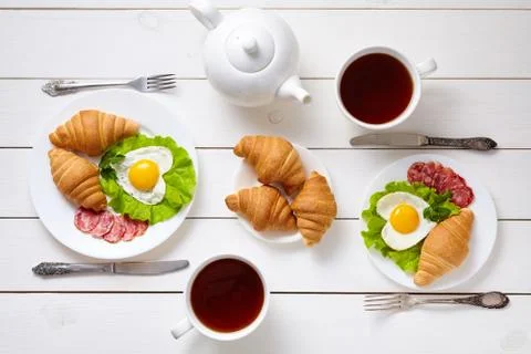 Heart shaped fried eggs, salad, croissants, salami sausage, composition and tea Stock Photos