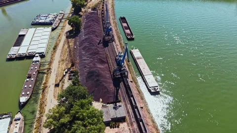 Heavy machinery loads cargo onto barges on a river bank surrounded by trees and Vídeos de archivo 344233269